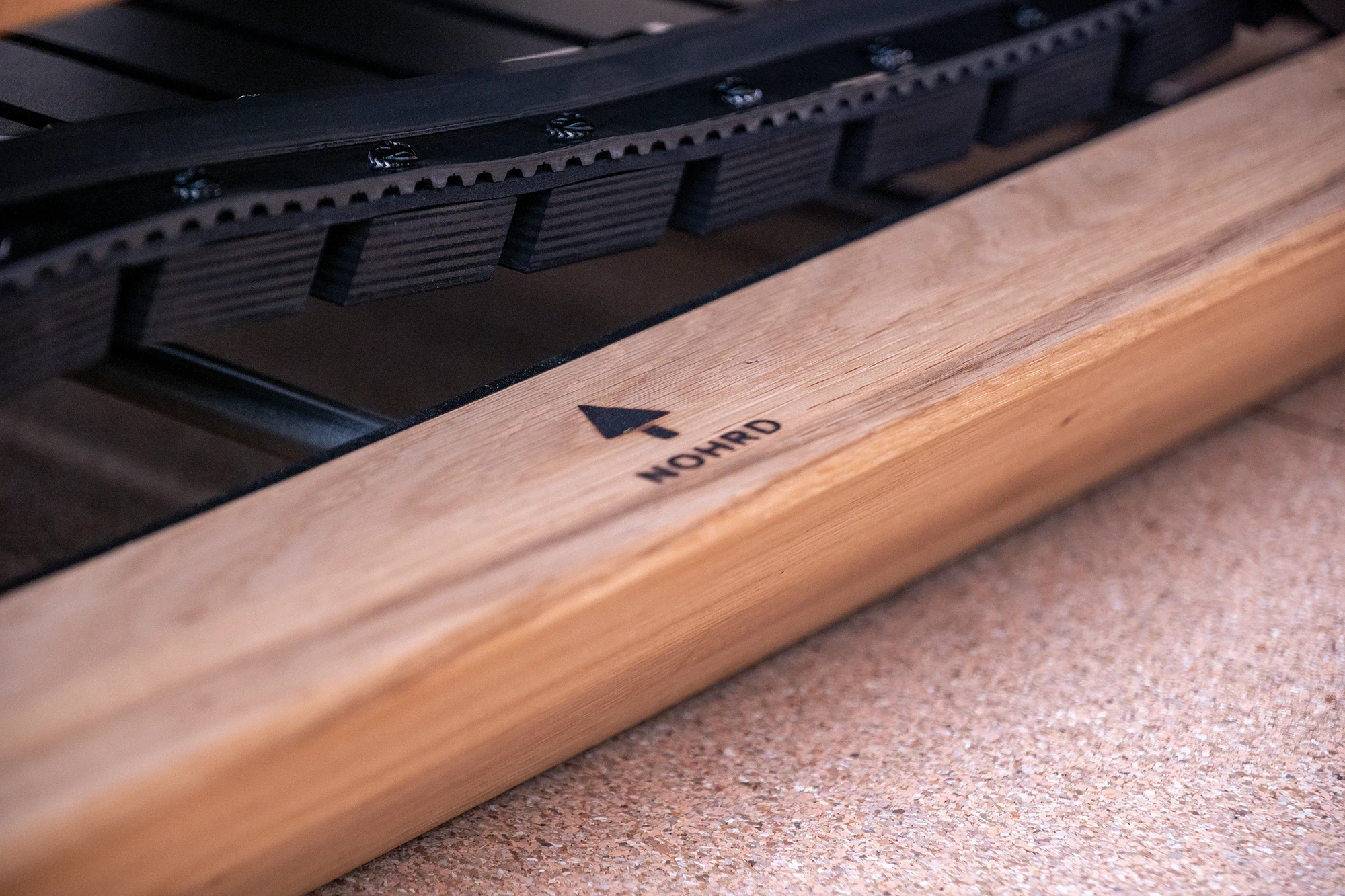 Détail d'un tapis de course en bois avec courroie crantée, flèche directionnelle et nom 'NOHRD' imprimé.
