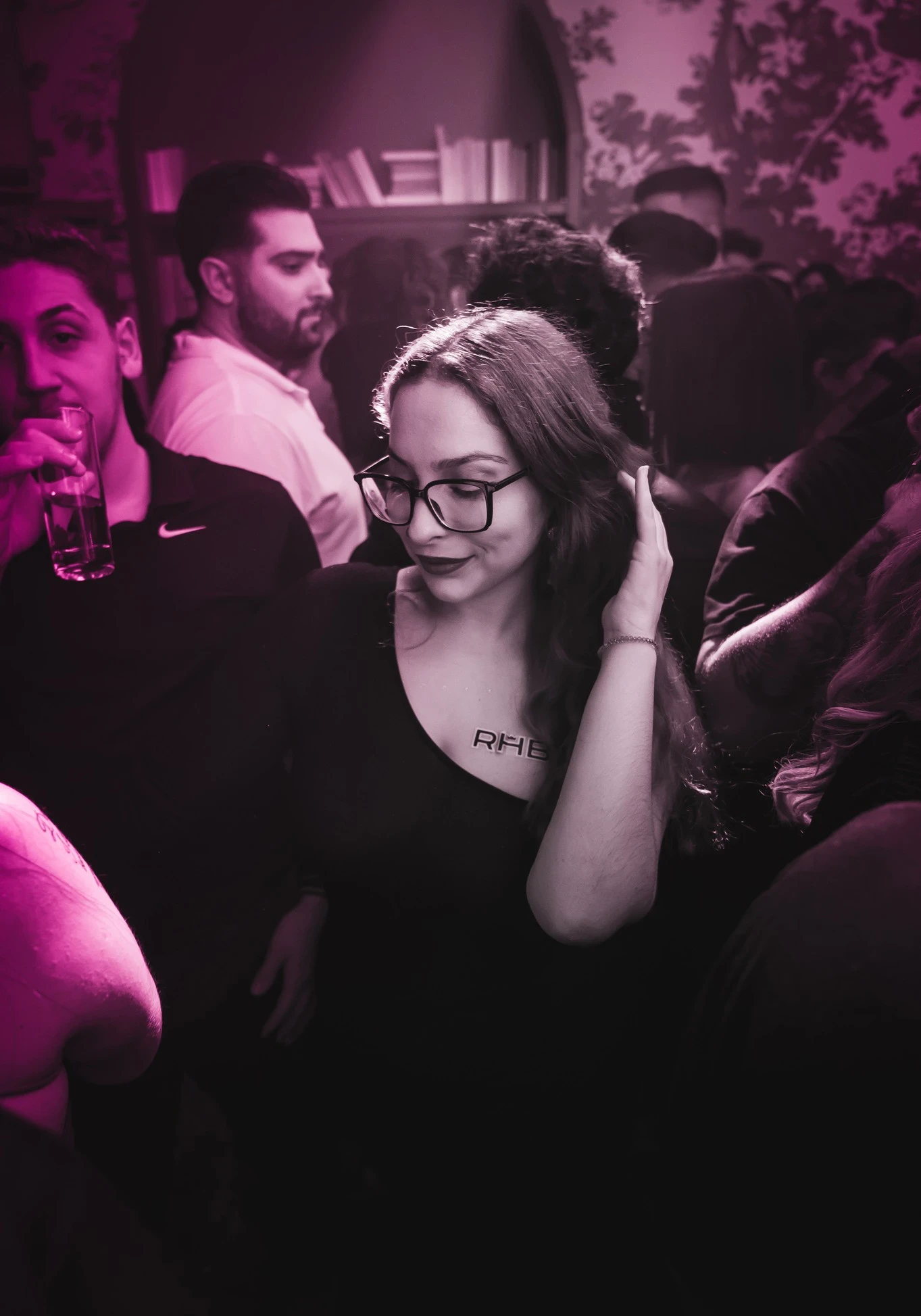 A person wearing glasses and a black top stands in the crowd at the RHB party. They are looking down and smiling slightly, with an "RHB" sticker on their chest.