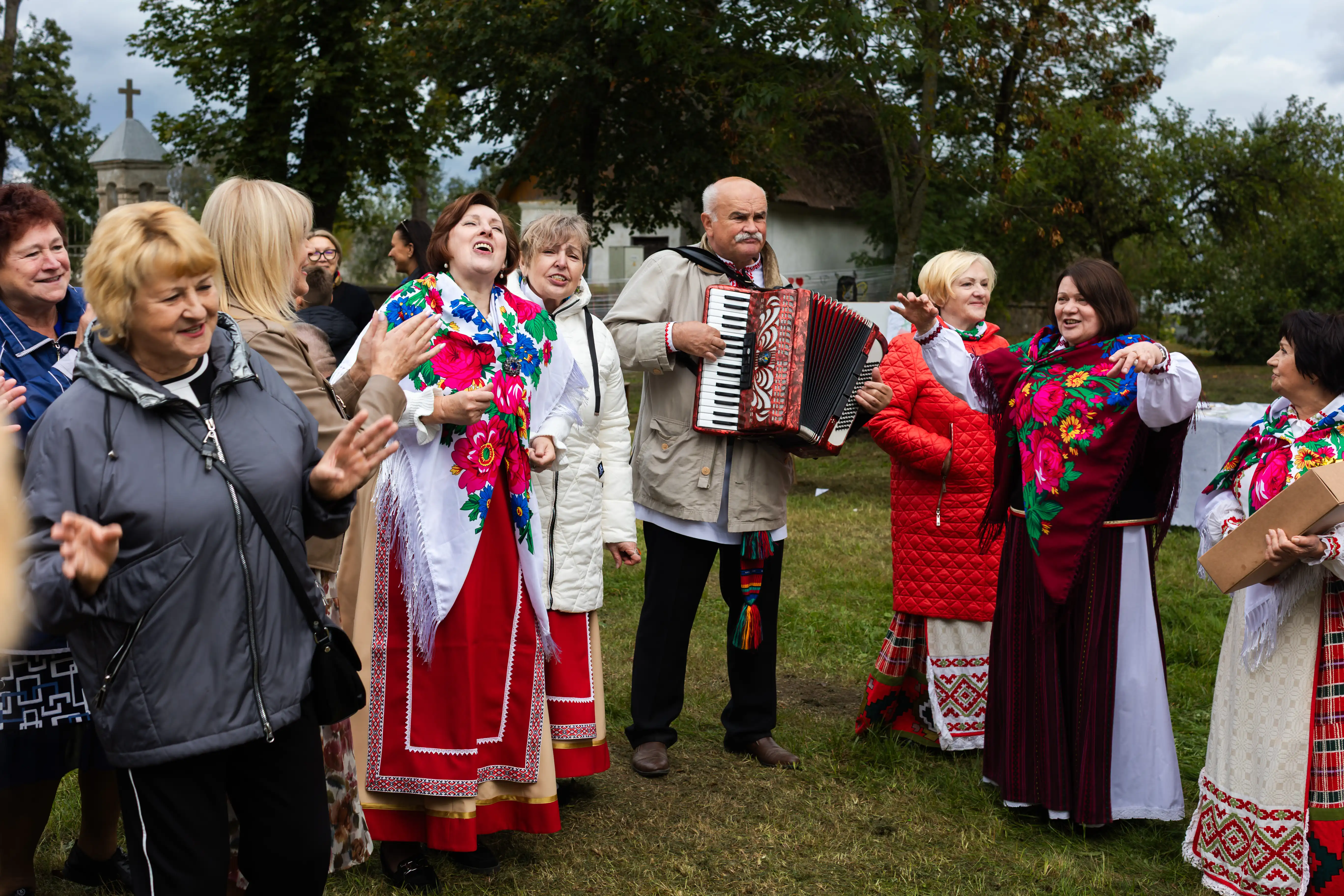 Grupa ludzi, głównie kobiet, tańczy i śpiewa na zewnątrz w tradycyjnych strojach. Mężczyzna gra na akordeonie, a w tle widać drzewa oraz budynek.