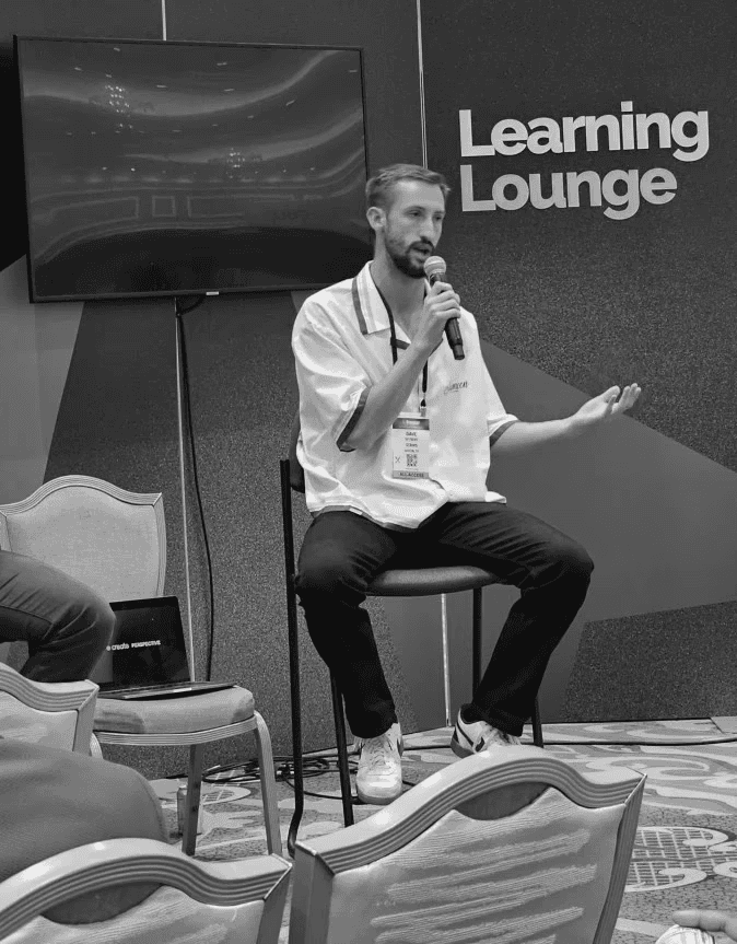 Black-and-white portrait of Thomas Layland standing in a modern office environment with blurred coworkers and plants in the background. He is wearing a light button-up shirt and smiling slightly with one hand in his pocket.