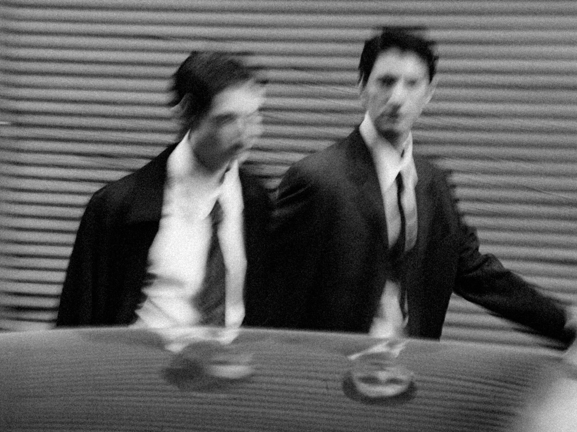 Blurred black and white photo of two men in suits walking past a corrugated metal background. Their indistinct features suggest motion and urgency.