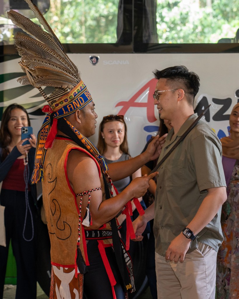 A traditional performer in ceremonial attire interacts with a visitor, while attendees observe the cultural exchange in a vibrant setting.
