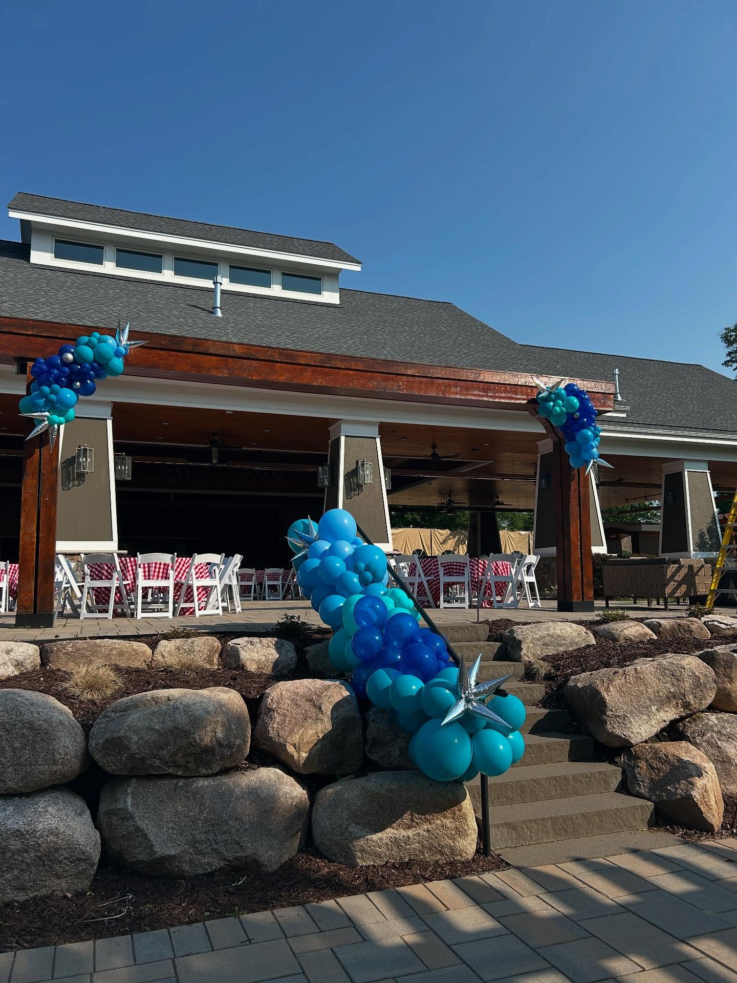 A festive outdoor event setup displays a pavilion with a sloped roof, adorned with vibrant blue balloon arches and clusters, in front of rock landscaping and neatly arranged tables and chairs inside.