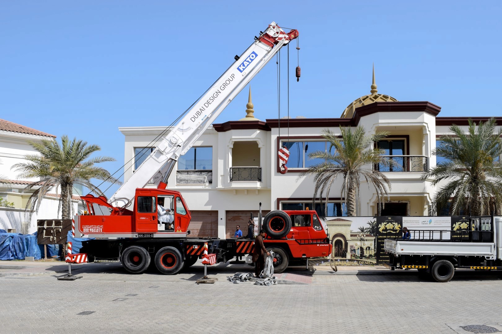 A large red industrial crane truck is positioned in front of a luxurious white villa with a golden dome, set against a clear blue sky, accompanied by palm trees and a parked black utility trailer on a paved driveway.