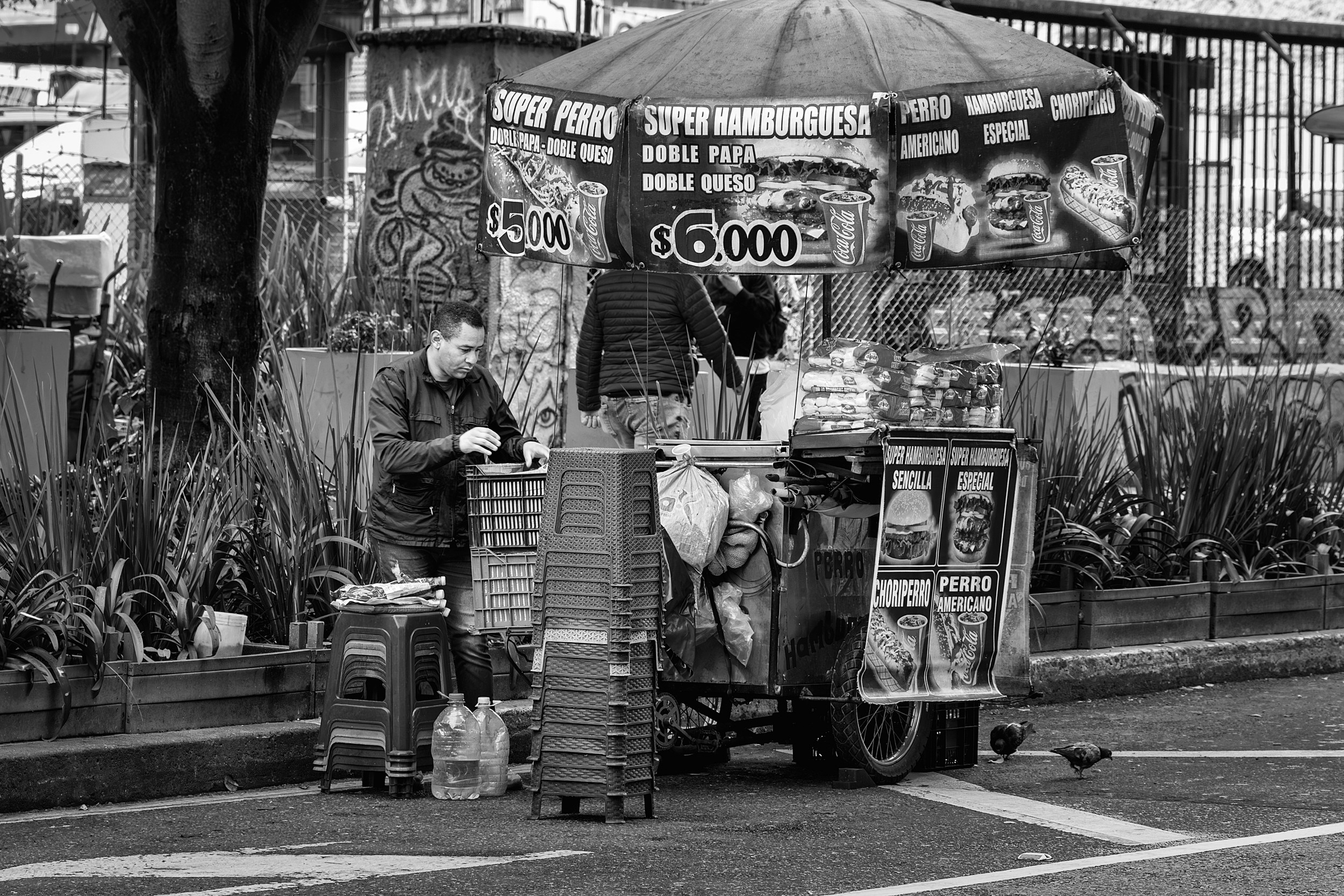 A street vendor prepares a food cart beneath a large umbrella advertising hamburgers and hot dogs in Bogotá, surrounded by plastic stools, crates, and urban greenery.