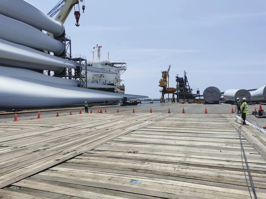 Port facility with offshore wind components, cranes, and workers operating on a timber quay.