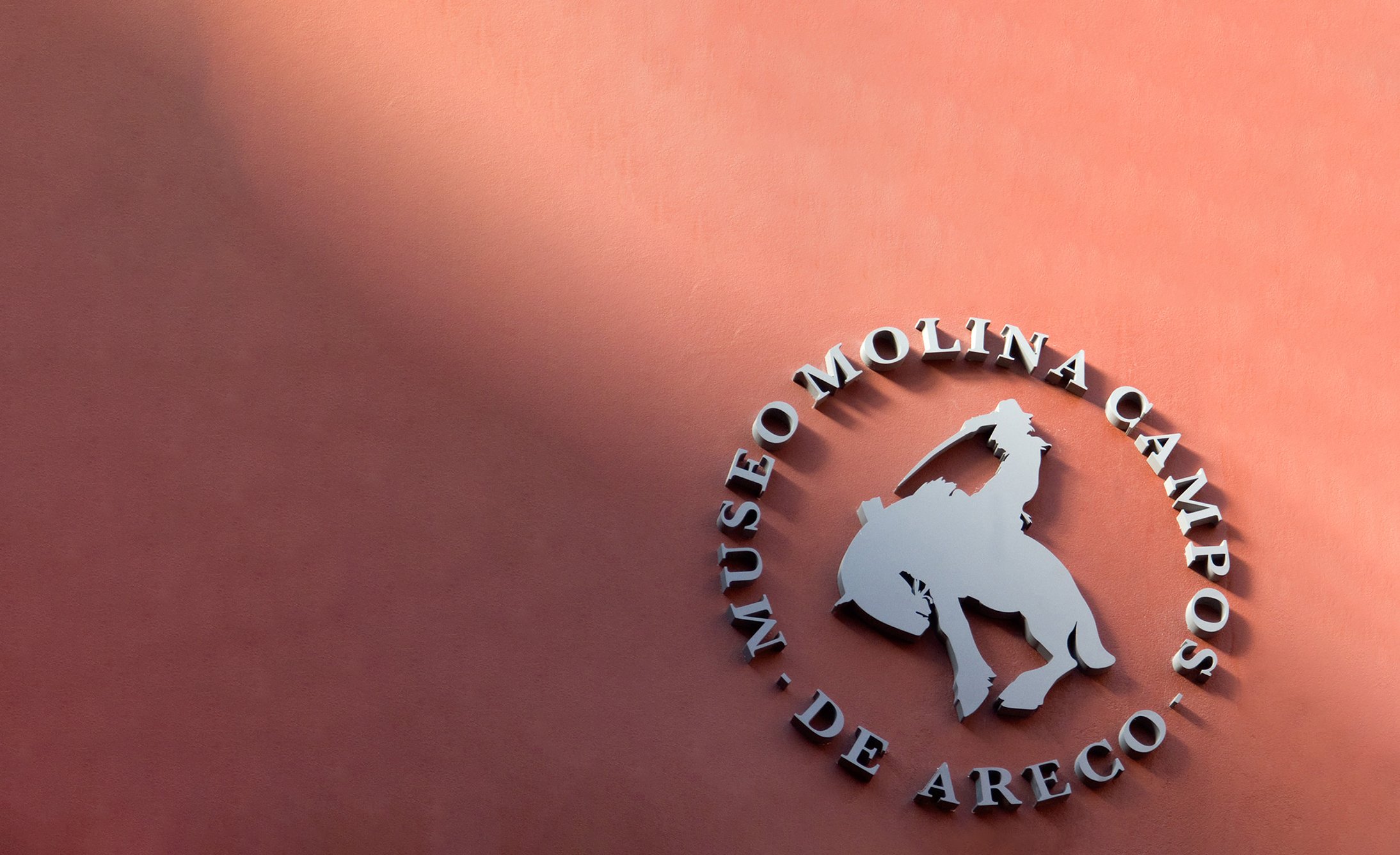 Metallic logo of Museo Molina Campos on a terracotta wall, featuring a silhouette of a cowboy on horseback under angled sunlight, casting shadows.
