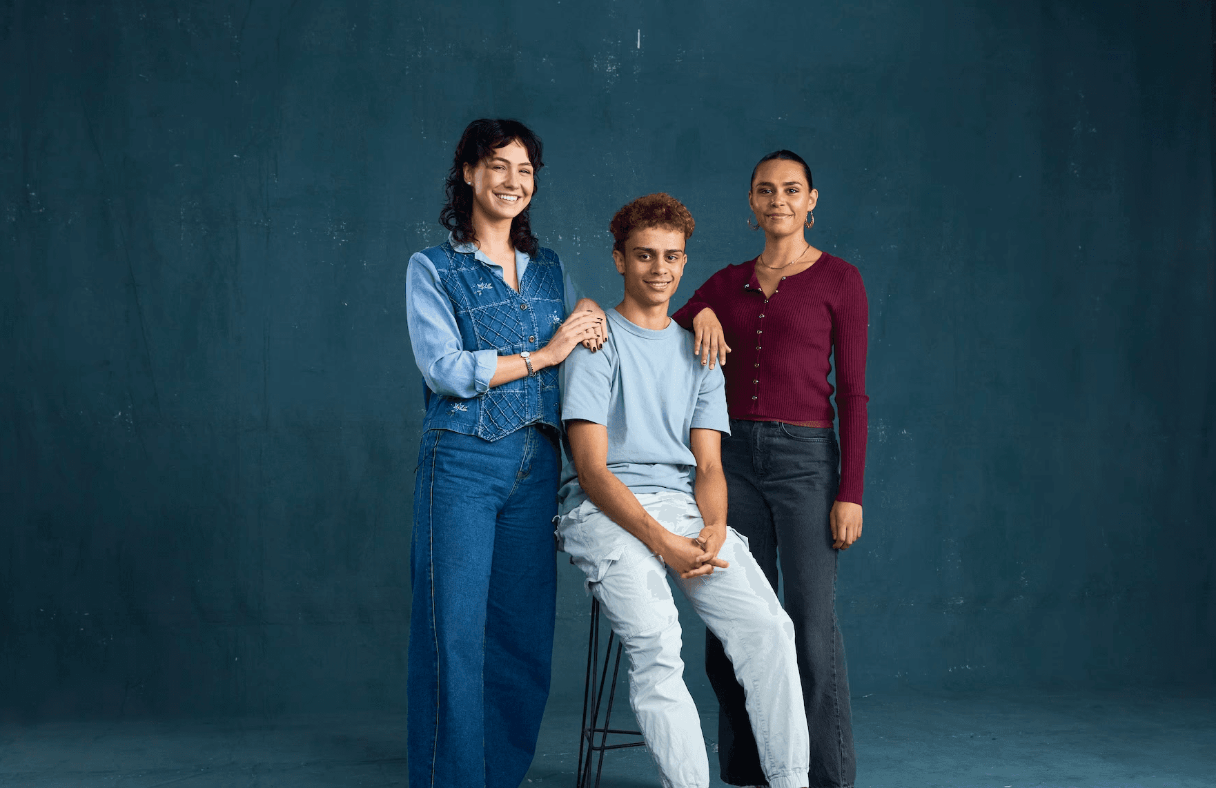 Three young adults pose together in front of a dark studio backdrop. One person sits on a stool wearing a light blue T-shirt, white pants and sneakers, while two others stand beside them with hands resting on their shoulders. The person on the left wears denim overalls and sandals, and the person on the right wears a maroon top and black jeans. All three look relaxed and smiling, with lighting equipment partly visible at the edge of the frame.