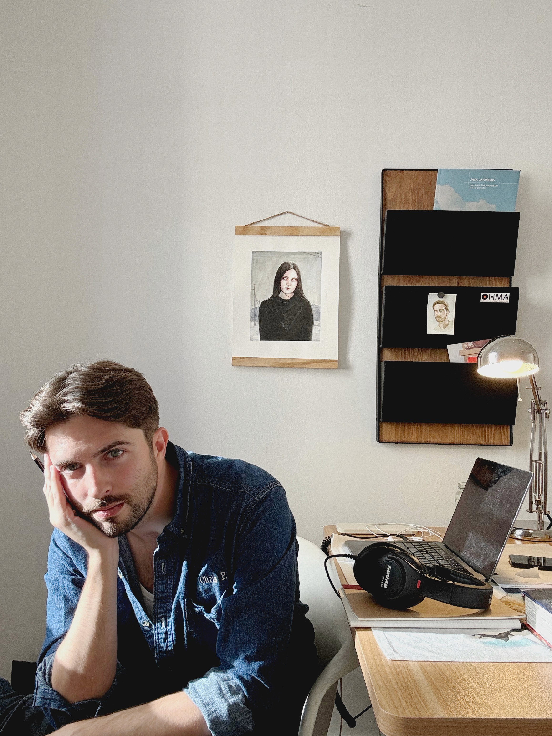 A man sits by a desk with his hand in his face. He has a painting, wall organizer, and laptop behind him.