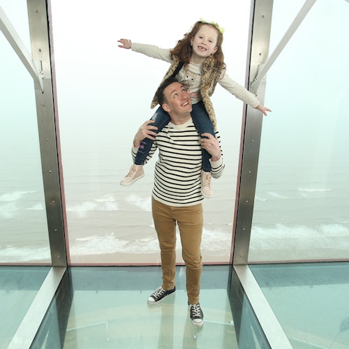 A man with a striped shirt holds a smiling girl on his shoulders, standing on a clear glass floor with a foggy background.