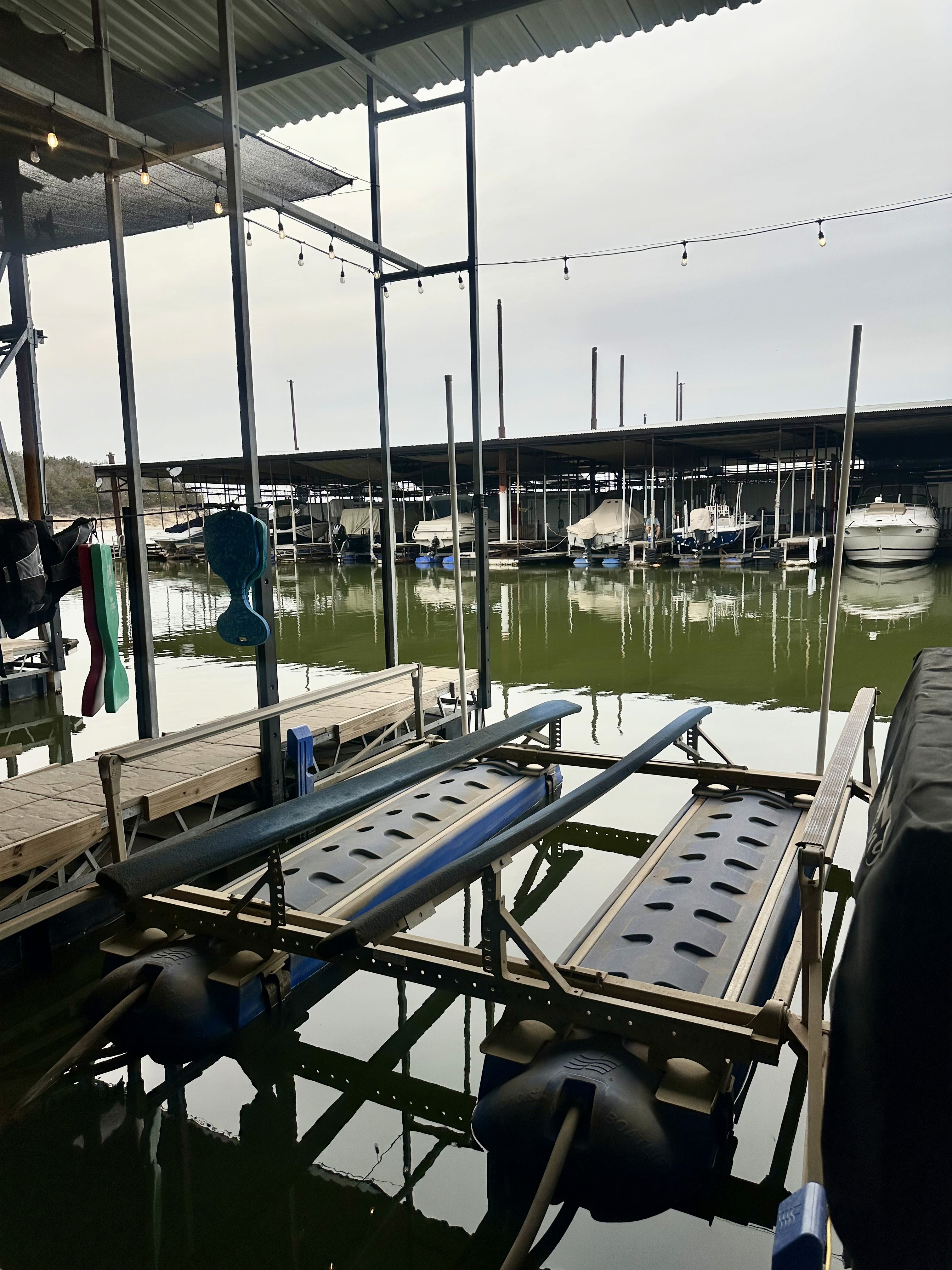 A tranquil marina scene featuring rows of docked boats under a large, covered structure with calm green water reflecting the overhead lights and surrounding architecture.