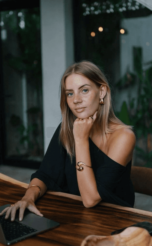 Young man in a black turtleneck and blazer stands outdoors, smiling softly. The background is blurred, conveying a warm, professional tone.