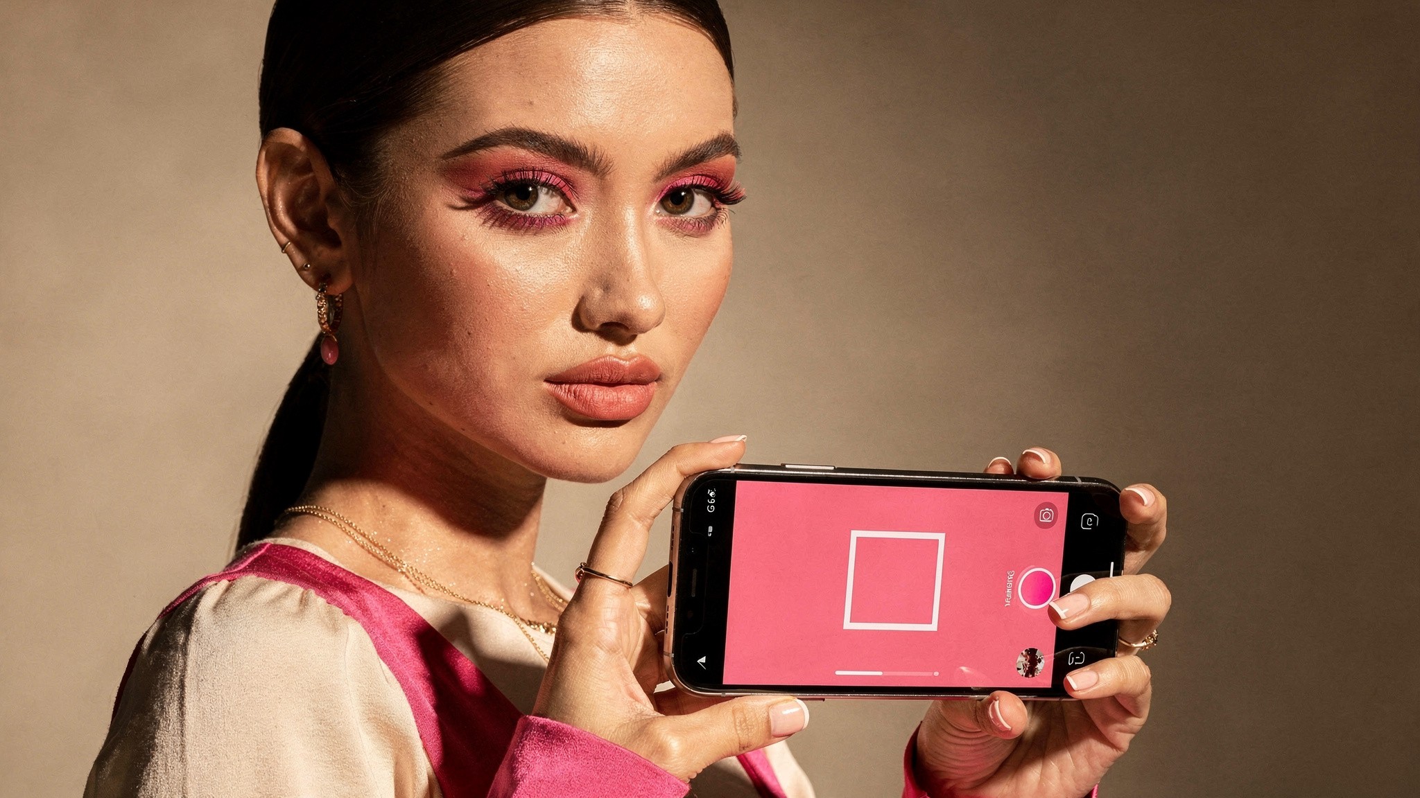 Beauty creator holding a phone showing a square Instagram photo in a pink studio