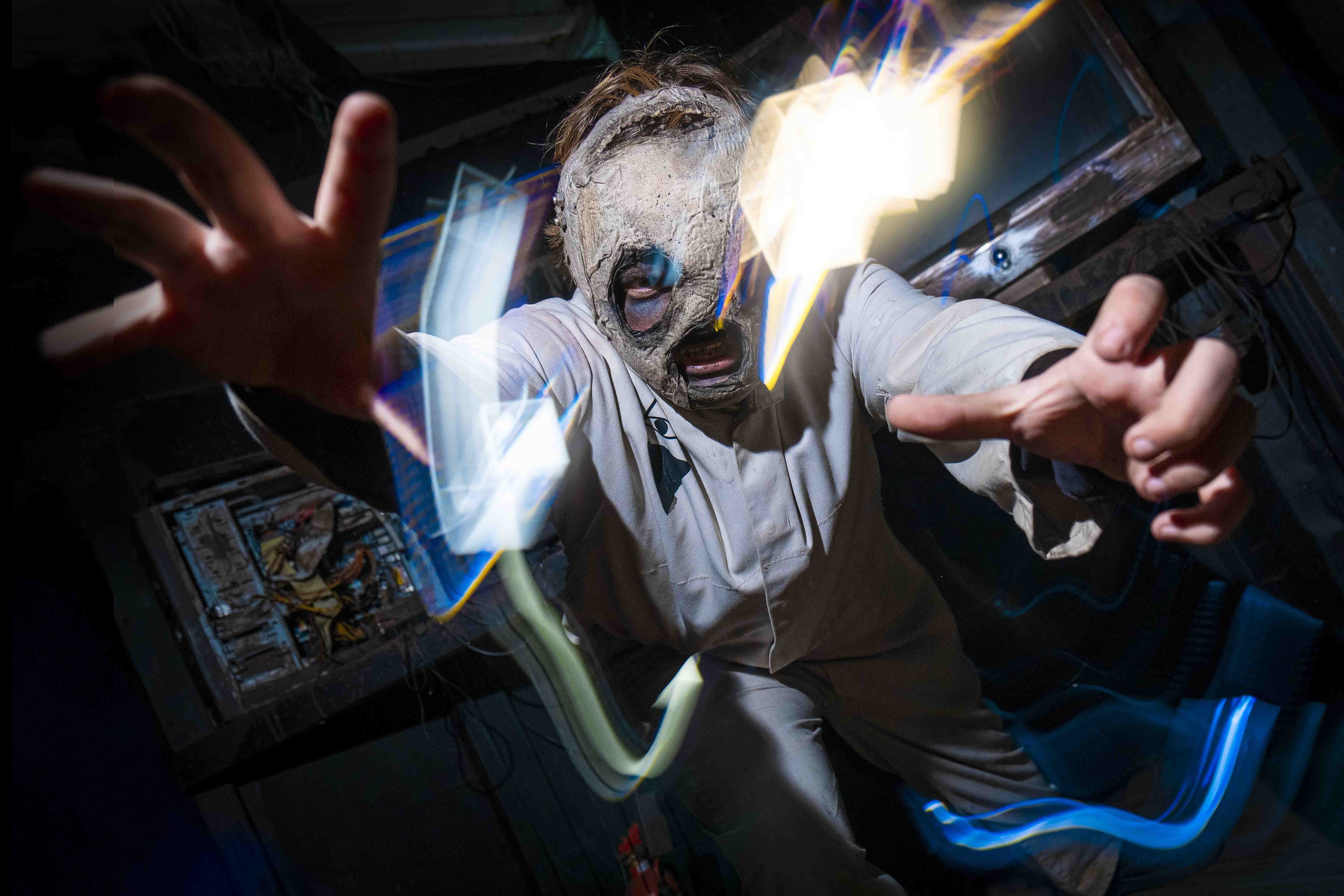 A person in a tattered mask and white clothing reaches forward menacingly, with colorful light streaks accentuating the eerie, dark setting, suggesting a horror or haunted theme.