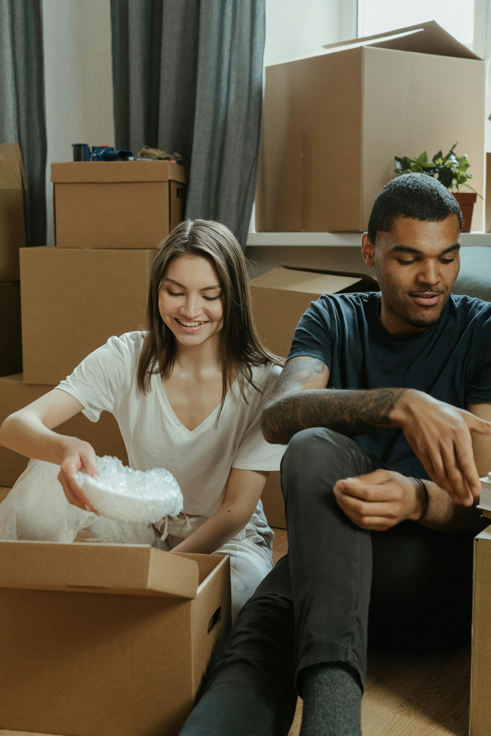 A couple is sitting on the floor surrounded by moving boxes. The woman, who has long brown hair, smiles while placing bubble wrap into a box. The man, who has tattooed arms, is looking down and reaching into a box next to her.