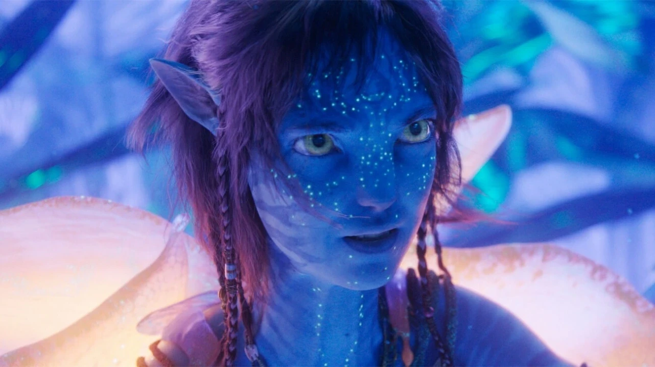 A young blue-skinned, otherworldly female figure with bright yellow eyes and luminescent dots on the face gazes intently in a vibrant, bioluminescent forest environment.
