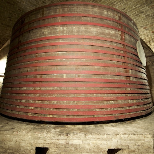 חבית יין גדולה מעץ עם רצועות מתכת אדומות במרתף עם קירות לבנים.