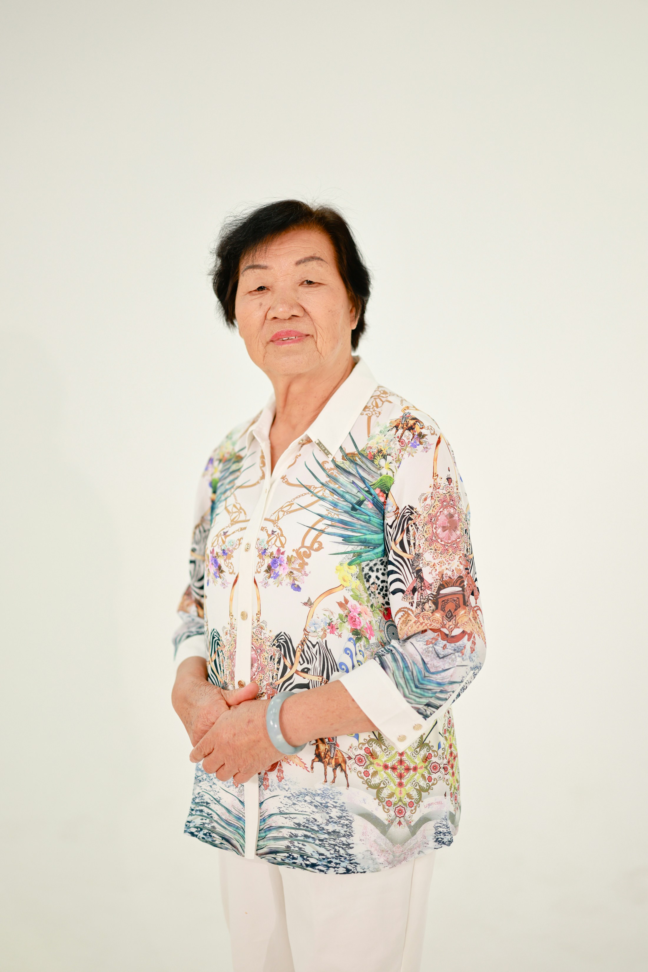 Elderly woman with a patterned shirt stands against white background.