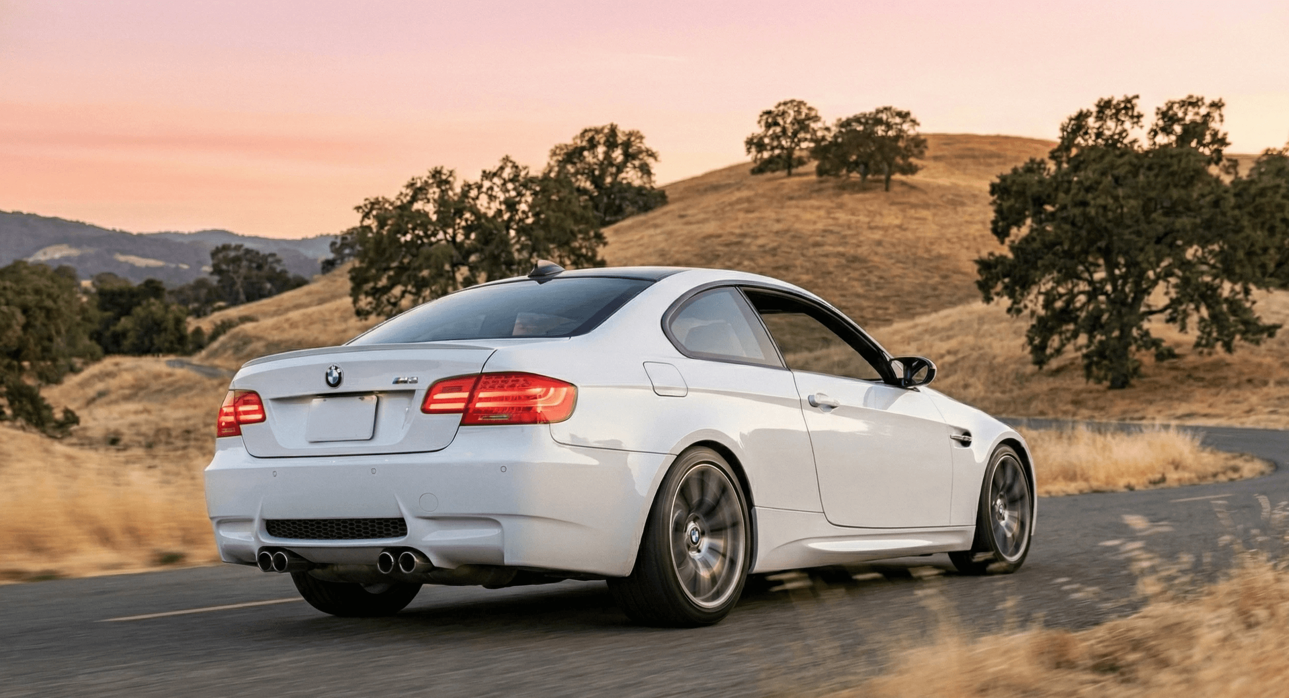 BMW M3 E92 in white accelerating on curved country road surrounded by oak trees and golden sunlight, representing documented enthusiast cars that command higher prices
