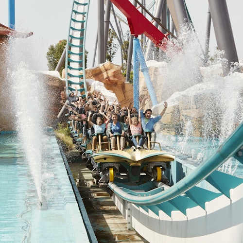 People riding a roller coaster with raised hands as it splashes through water, creating large water sprays.