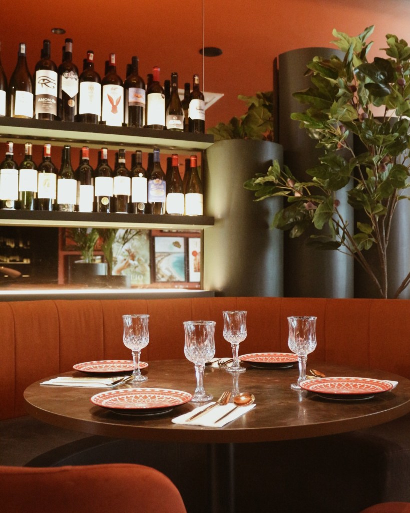 A dining table set with elegant glassware and decorative plates, surrounded by a plush red booth and lush greenery, evoking a warm and inviting atmosphere.