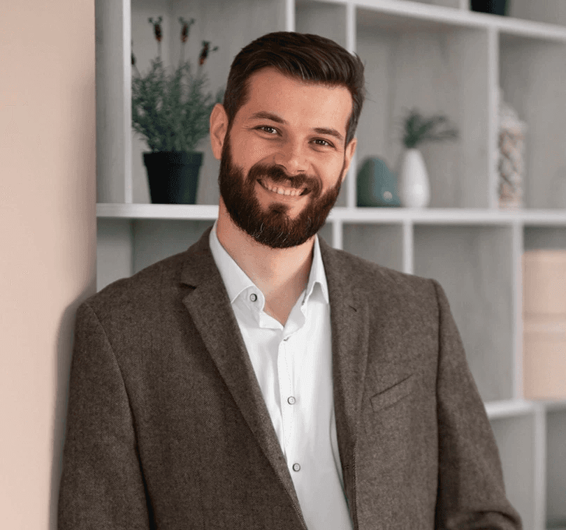 Smiling man with a beard, in a brown jacket, leans against a wall holding a laptop. Behind him is a shelf with plants and decorative items.