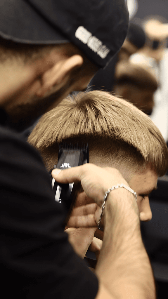 A barber is using clippers to style a client's hair in a barbershop setting.