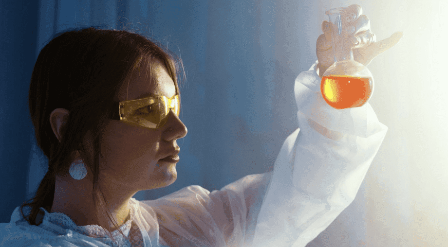 A young female scientist examining a flask with orange liquid in a laboratory setting. She wears protective golden goggles and a white lab coat, emphasizing safety in her work environment.