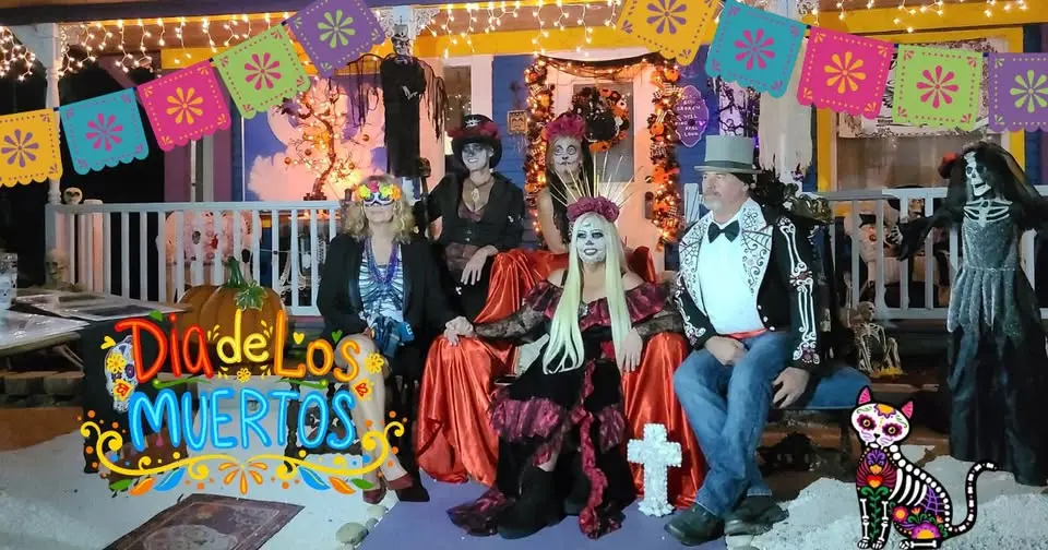 A group of people in elaborate Dia de los Muertos costumes are seated in front of a brightly decorated residence in Bradenton, FL, featuring colorful papel picado banners and themed decorations like skeletons and marigolds.