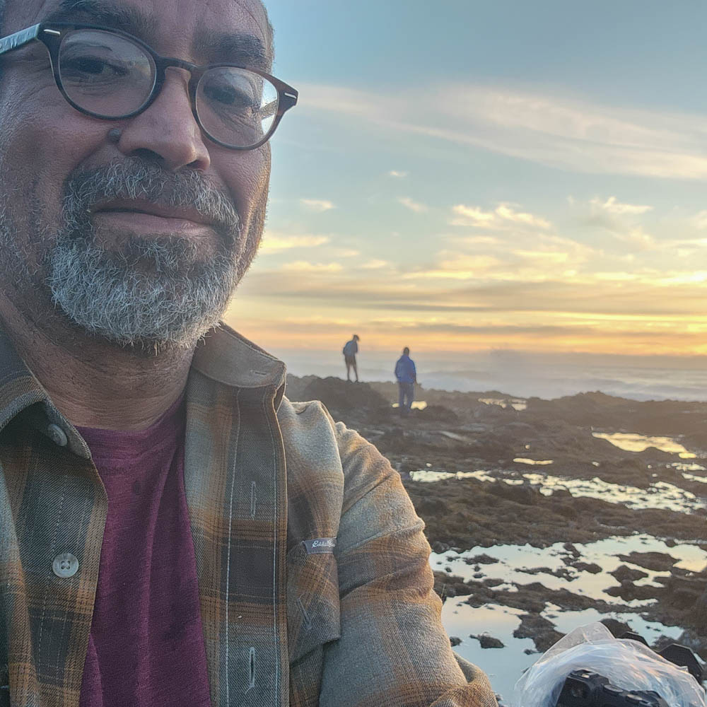  Benjamin taking a selfie near rocky tidepools at sunset with two people walking in the distance.