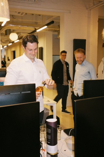 photo of the man opening a bottle of alcohol and looking into it. Two men on the background looking for the one with.a bottle. Office background and friendly atmospere