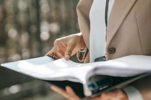 Businesswoman in tan blazer reviews documents, holding file. Office professional reviewing business paperwork.
