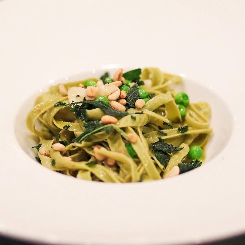Gourmet green ribbon pasta dish featuring fresh garden peas, toasted pine nuts, and crispy sage leaves served in a minimalist white ceramic bowl.