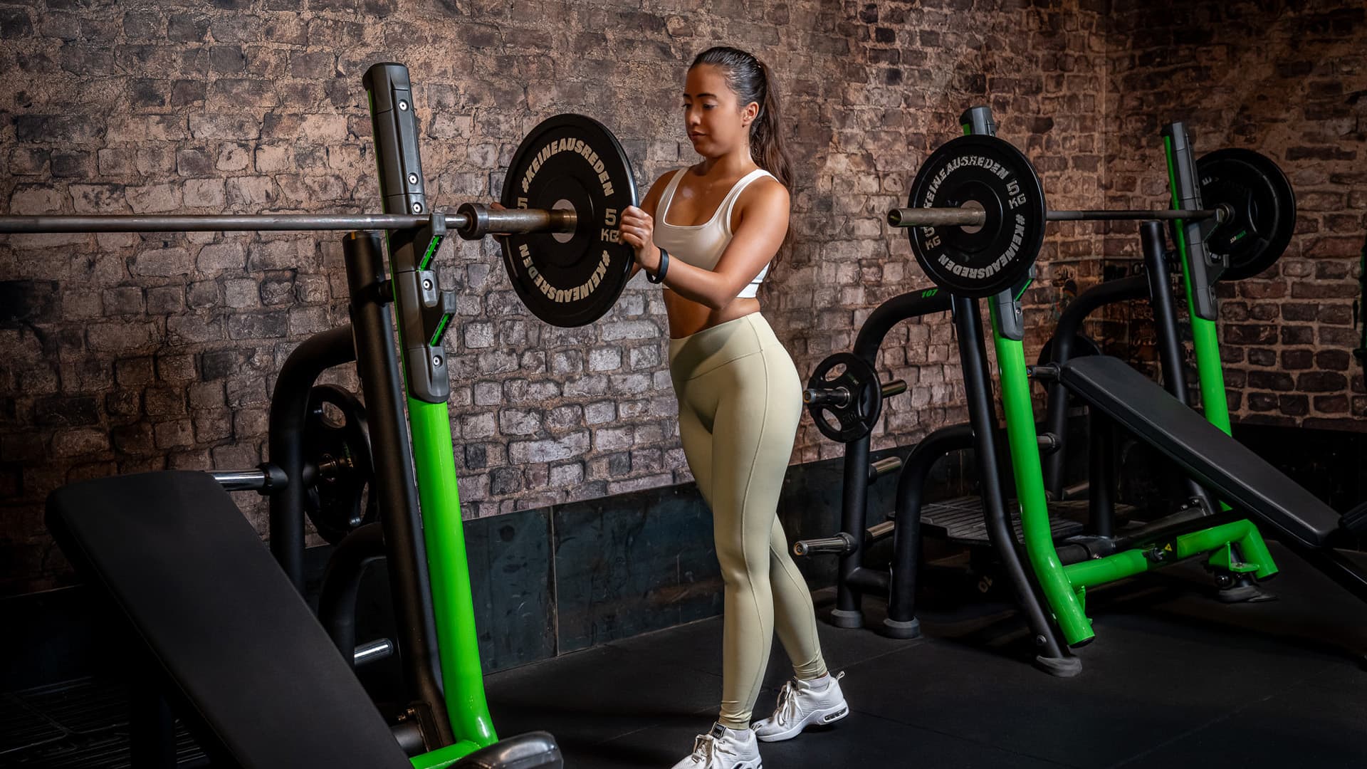 Frau legt Gewichtsscheibe an die Langhantel im Fitnessstudio und steigert ihr Trainingsgewicht