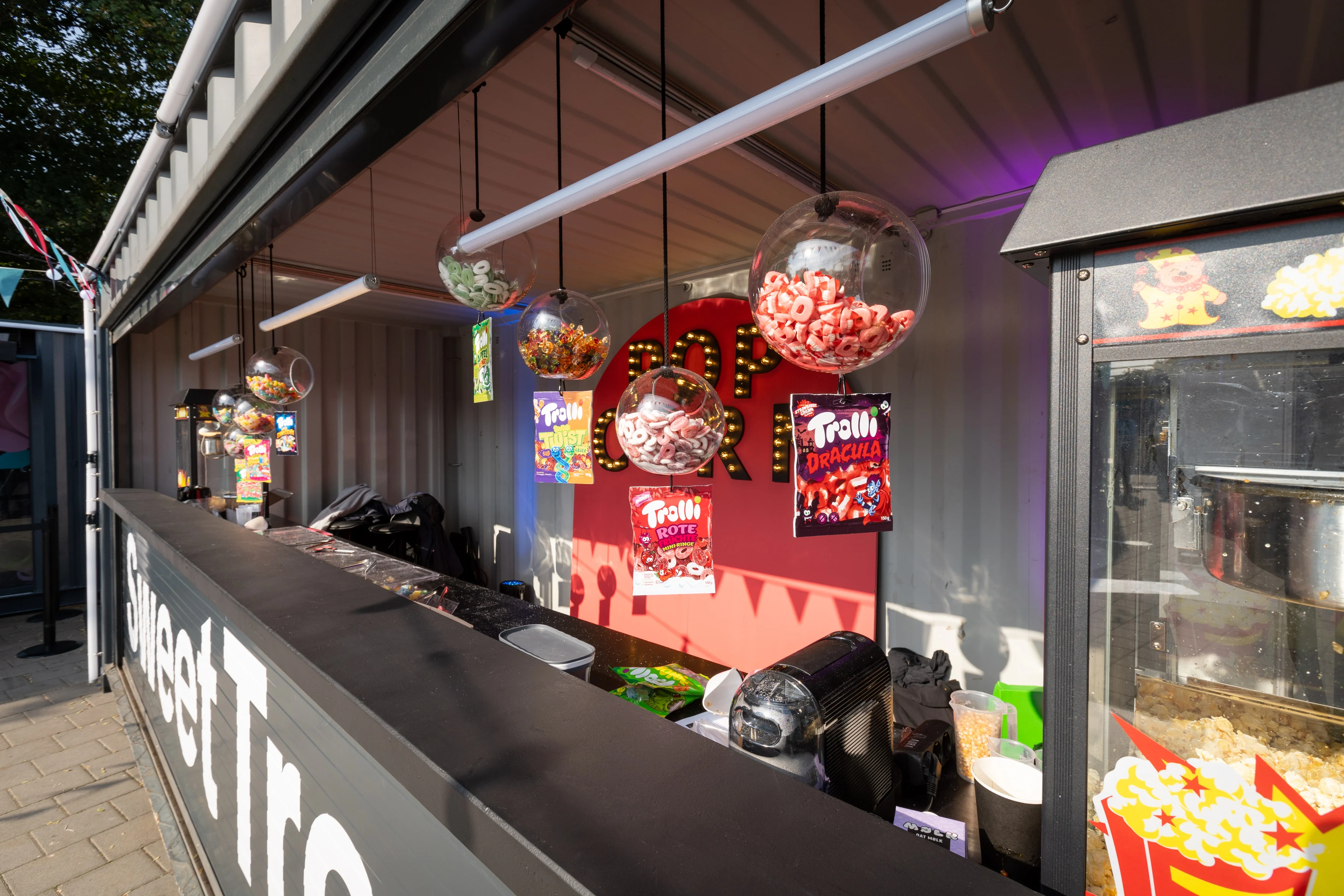 Sweet treats counter in a container with hanging candy spheres, popcorn machine and Trolli signs.