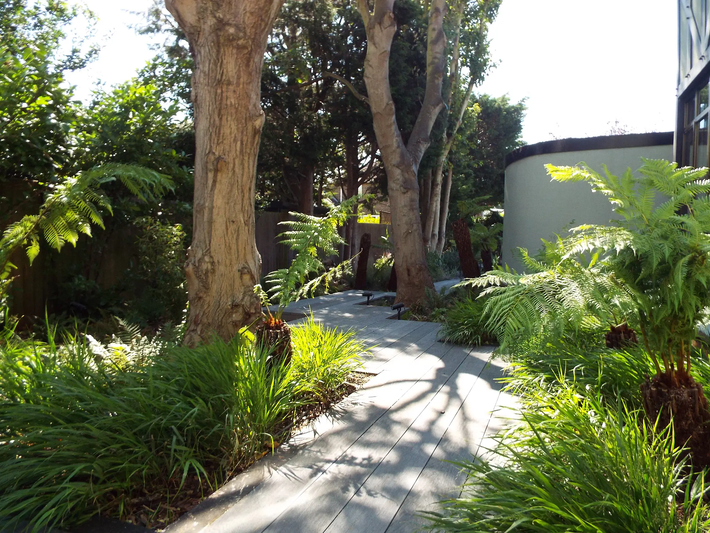 A sunny pathway lined with greenery, leading towards trees and a building in a lush garden setting.