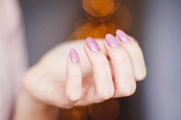A hand with almond-shaped nails in dusty pink gel polish, one nail in pink glitter, against soft bokeh lights