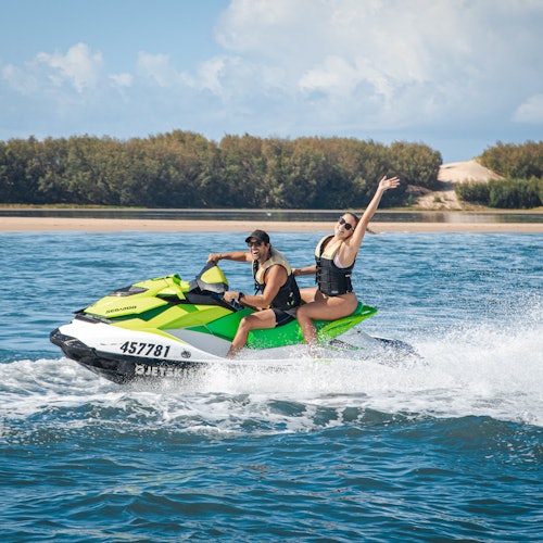 Two people riding a green jet ski in open water, one raising an arm. A distant sandy shore with trees is visible in the background.