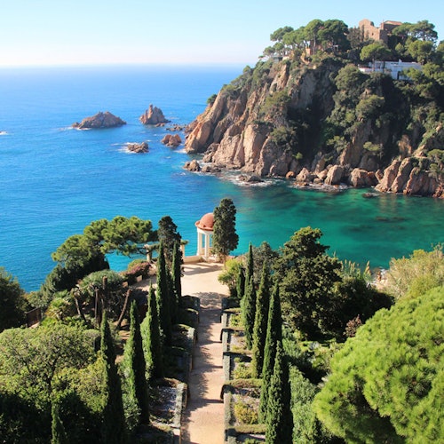 地中海沿岸景色，礁石、碧绿色海水，以及一条由柏树环绕的花园小径通向一个小凉亭。