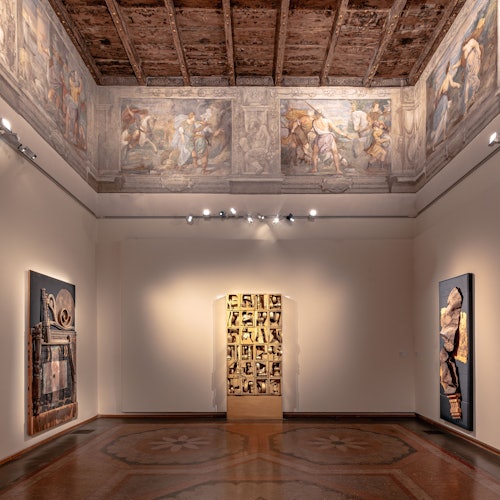 Sala della galleria d'arte con soffitto affrescato, opere d'arte moderne alle pareti e un ingresso ricoperto di foto sul retro.