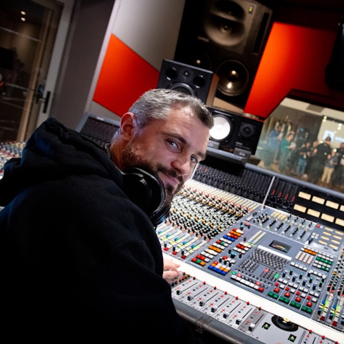 A man with headphones around his neck in a recording studio, sitting at a large mixing console.