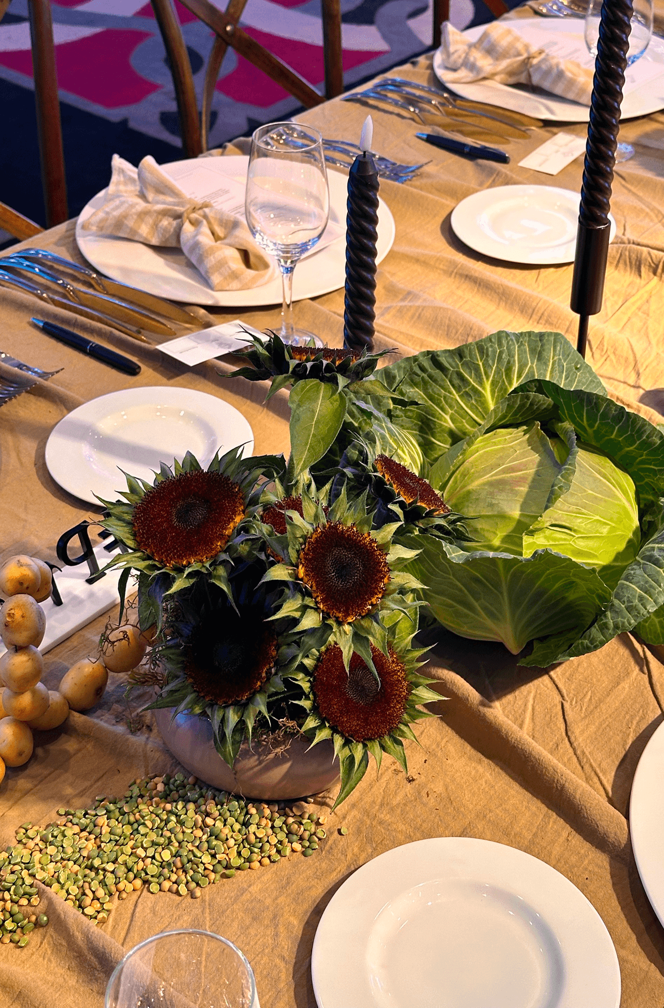 Decoración rústica con girasoles, repollo natural, velas negras y legumbres secas sobre mantel de lino beige.