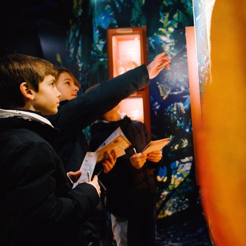 Tři děti v bundách se soustředěně dívají a ukazují na osvětlený displej, přičemž drží v rukou brožury.