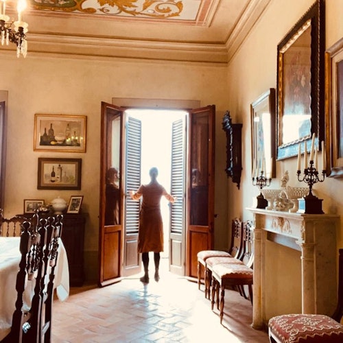 一个人站在一个精美装饰的房间里，打开门迎接明亮的阳光。房间里有艺术品、椅子和一个壁炉。