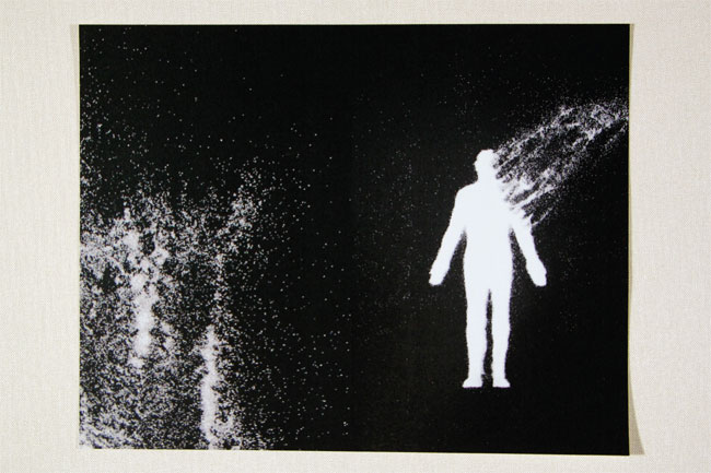 A white silhouette of a person stands against a black background. The figure is positioned slightly off-center, facing right. A diagonal white streak extends from the figure’s right shoulder, curving upward and to the right. The left side of the image is filled with small, white specks, resembling a starry or speckled effect. The edges of the image are framed by a light-colored border.