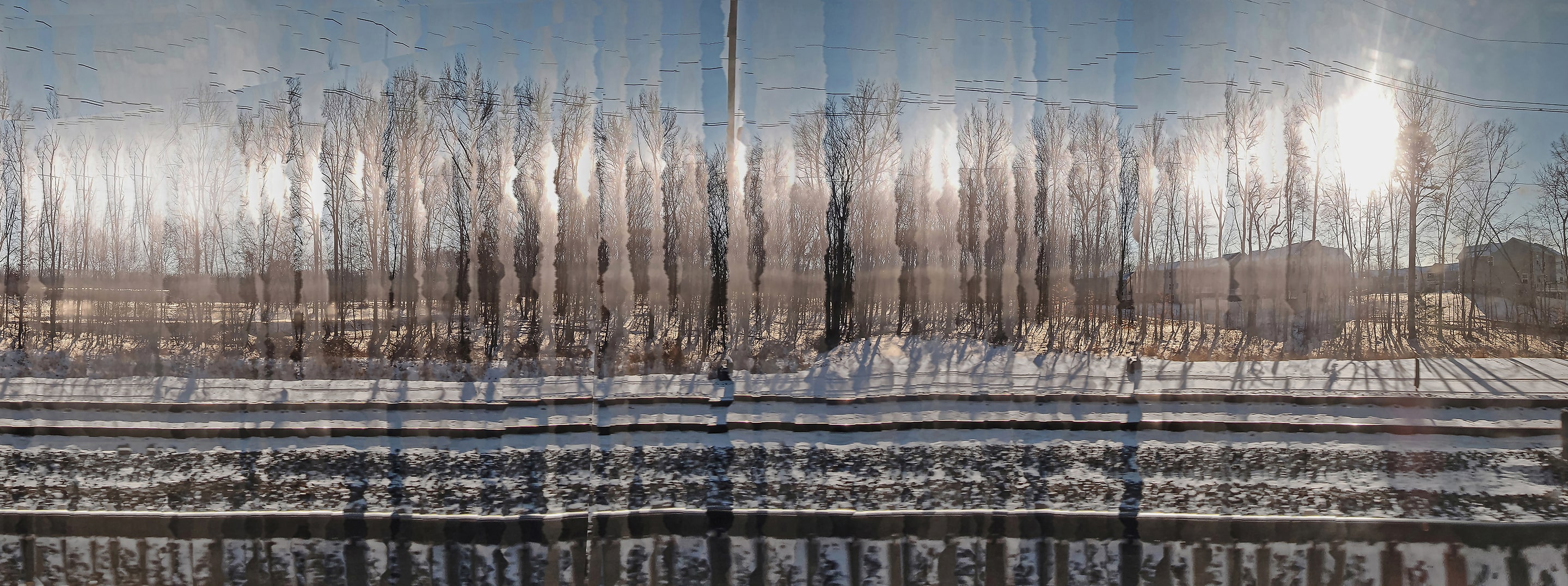 Panoramic photograph of leafless trees along snowy train tracks, captured through a distorted motion effect that stretches light and shadow into vertical bands. The image blurs the line between landscape and time, evoking memory and passage.