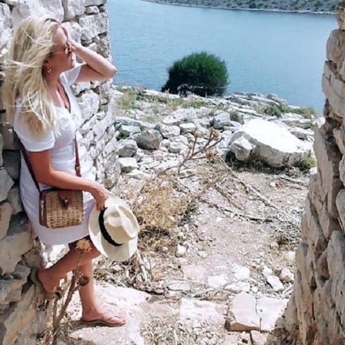 En kvinna i en vit klänning och sandaler står mellan stenmurarna, håller en hatt och blickar ut över en sjö och ett klippigt landskap.