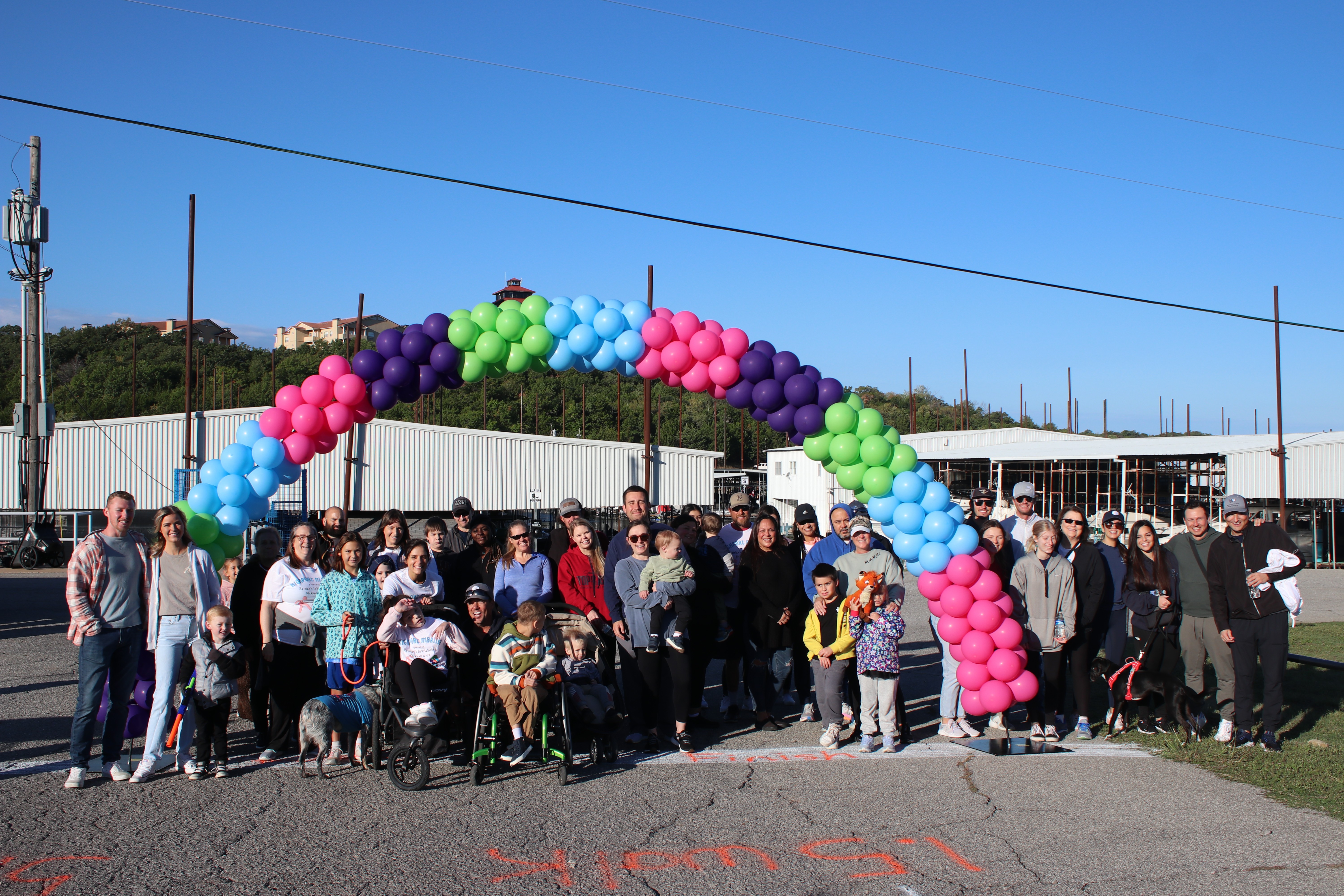 A large, diverse group of people, including children and a participant in a wheelchair, gather outdoors under a vibrant balloon arch made of pink, purple, green, and blue balloons, with industrial buildings and a clear blue sky in the background.