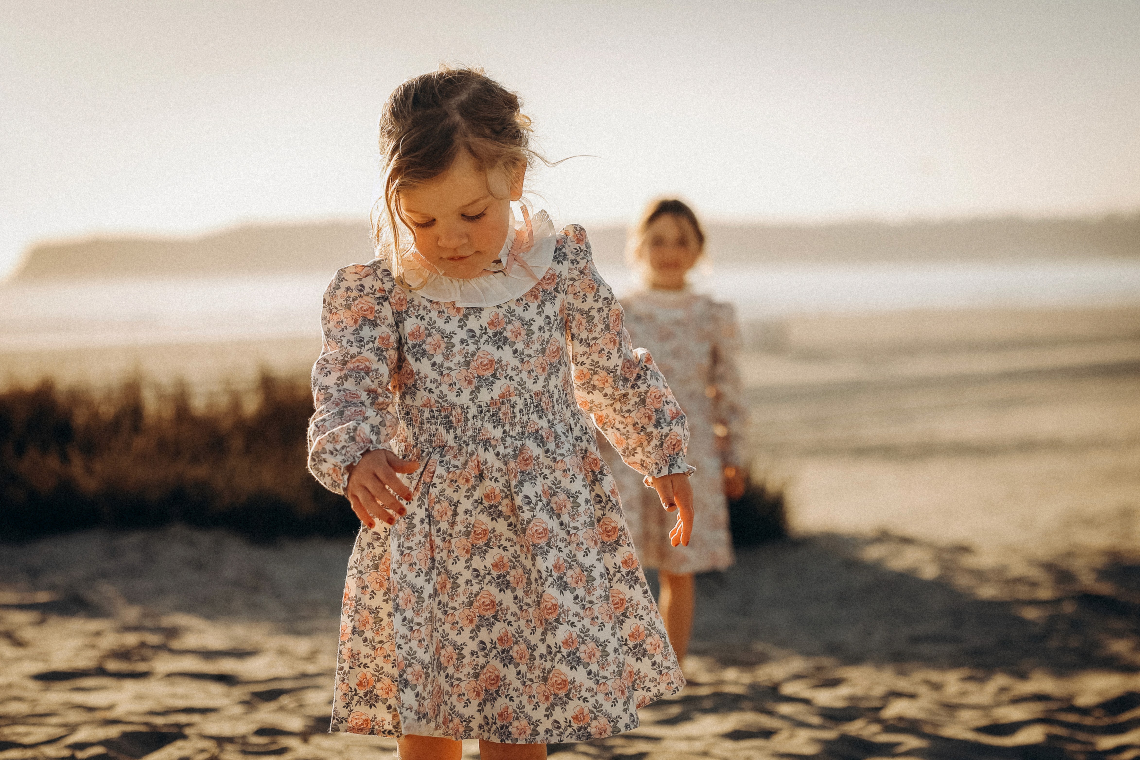 Little girl walking toward the camera with her sibling playing behind her on the beach.