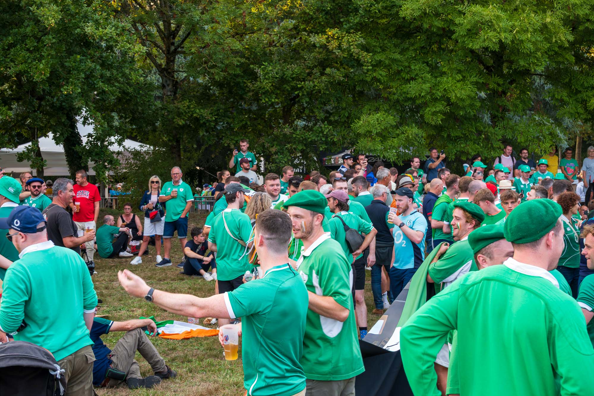 Fan zone de la Beaujoire avant Irlande – Tonga : foule de supporters irlandais en maillots verts et bérets verts sous les arbres, pintes à la main, drapeau irlandais posé sur l’herbe